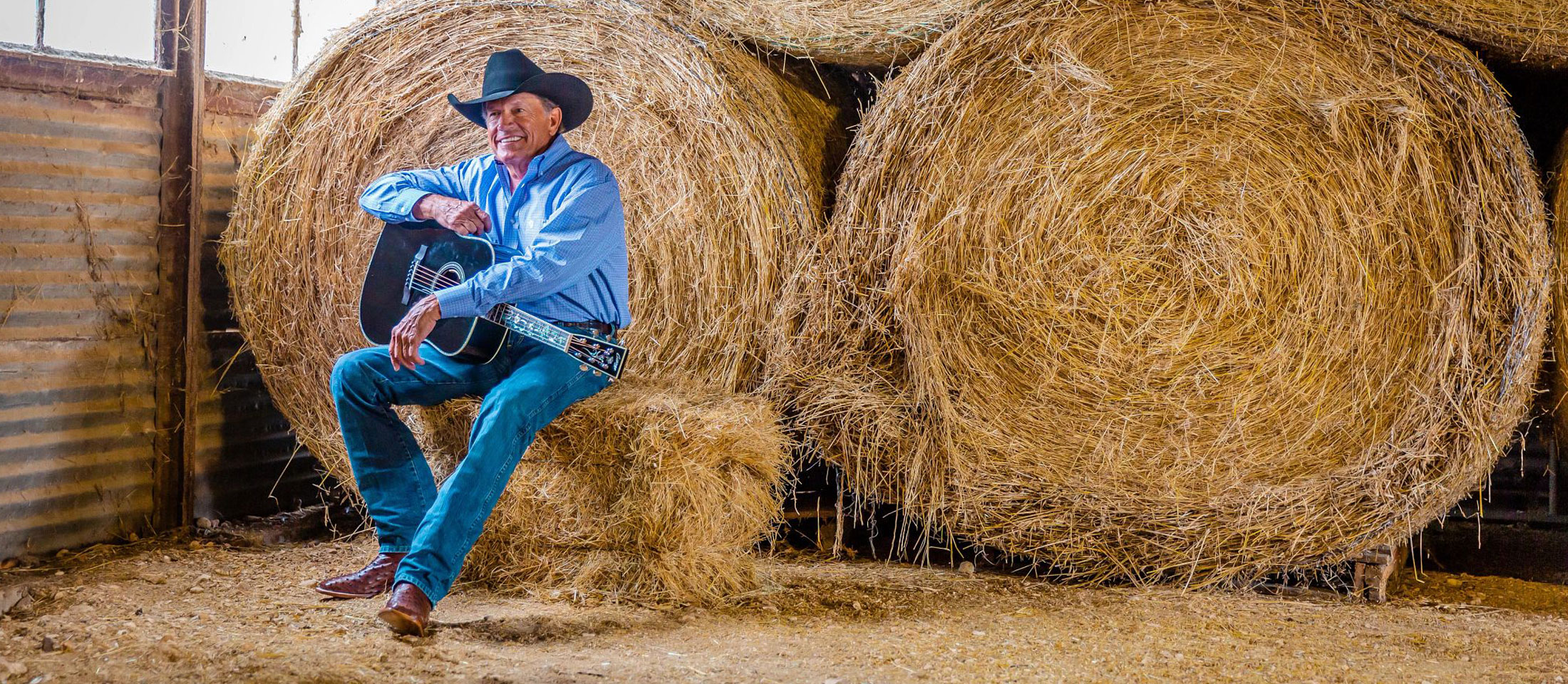 George Strait sitting on a square bale holding a guitar smiling.